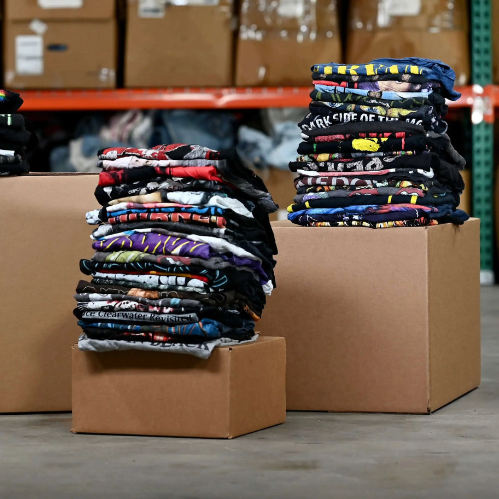 Stacks of vintage-style t-shirts on cardboard boxes in warehouse – part of a 15-pack mystery tee bundle.